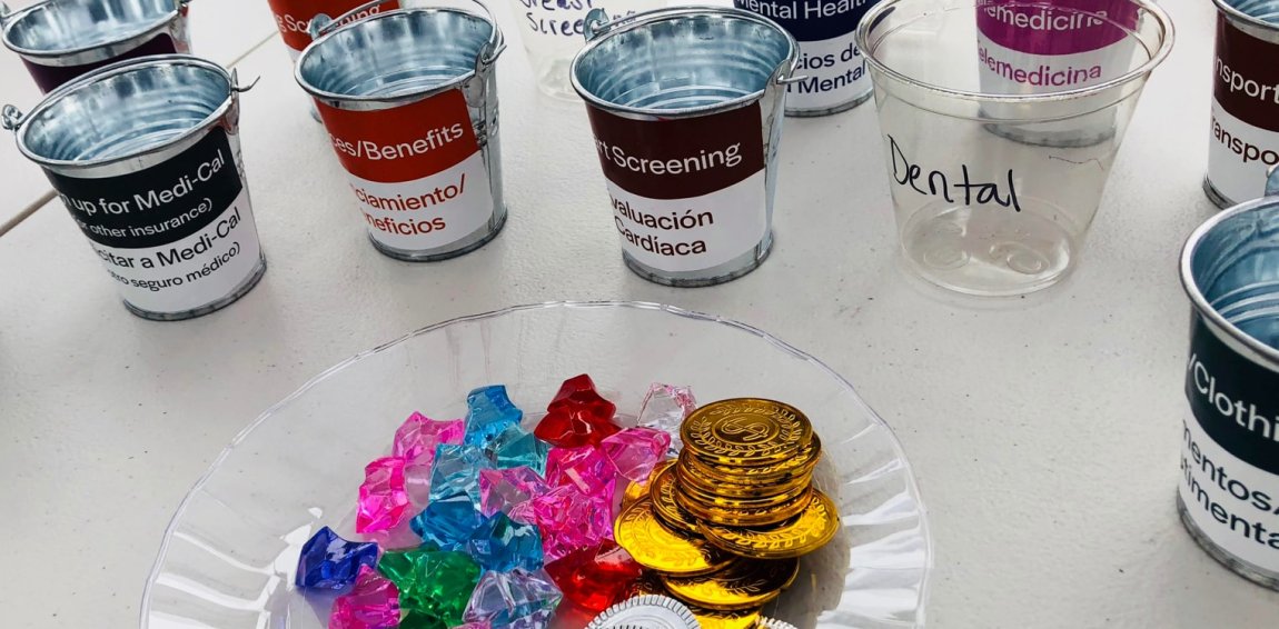 Piles of toy jewels, gold and silver coins sit near empty cans labeled with health-related needs such as mental health, signing up for Medi-Cal, and health screening